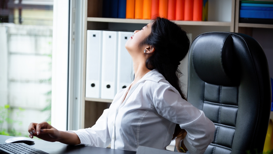 久坐办公室女性出现背部与下半身不适情境示意图，佳思优医美整形诊所大肠直肠外科医师赖依伶提醒久坐可能增加痔疮风险